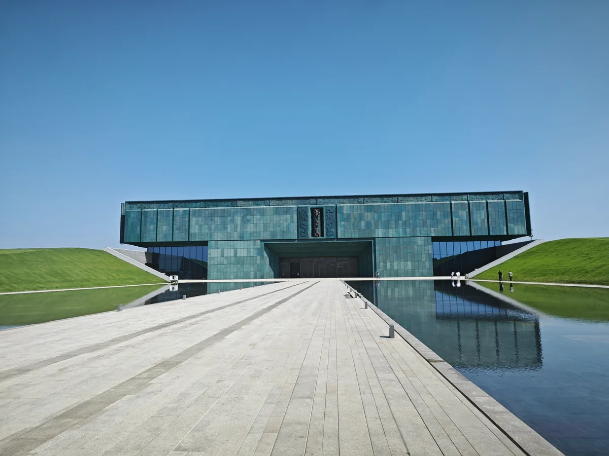 O Museu de Yin Xu em Anyang, um edifício baixo e comprido revestido de painéis verde-bronze, refletido num canal de água raso sobre um largo pavimentado (Photo: 1969社论 via Wikimedia Commons)