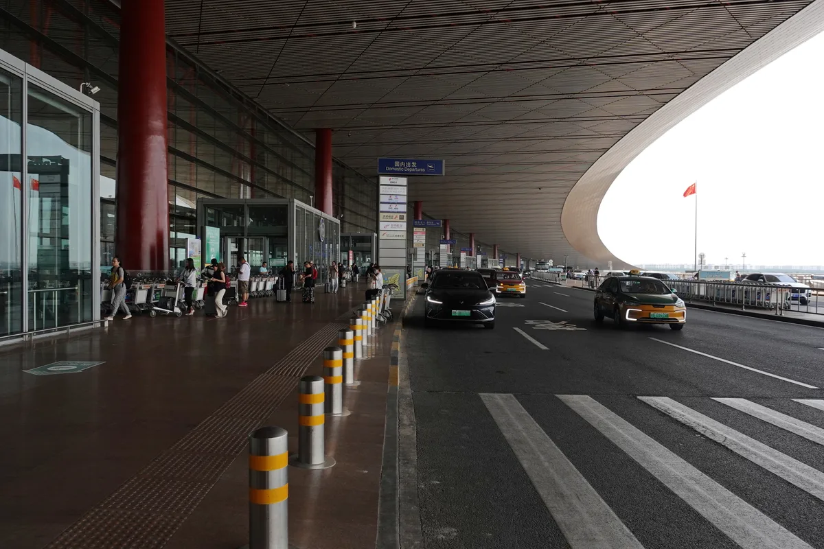 The departures curb at Beijing Capital Airport Terminal 3, with red columns, taxis and passengers wheeling luggage (Photo: 颐园居 via Wikimedia Commons)