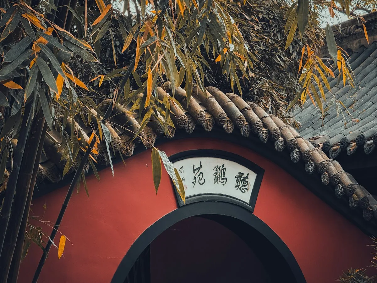 Un muro de jardín rojo con tejado de tejas grises y un arco bajo una placa blanca de caligrafía china, enmarcado por bambú