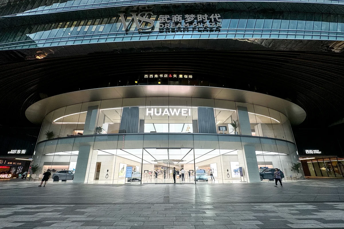 The curved glass frontage of a Huawei store at the WS Dream Plaza shopping mall in Wuhan at night, with cars on display inside and shoppers on the plaza outside