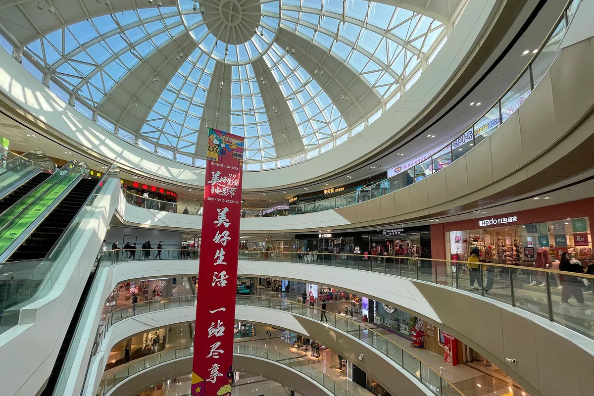 The glass-domed atrium of a shopping mall in Zhengzhou, with escalators, curved balconies and a long red banner hanging through the open space (Photo: Windmemories via Wikimedia Commons)