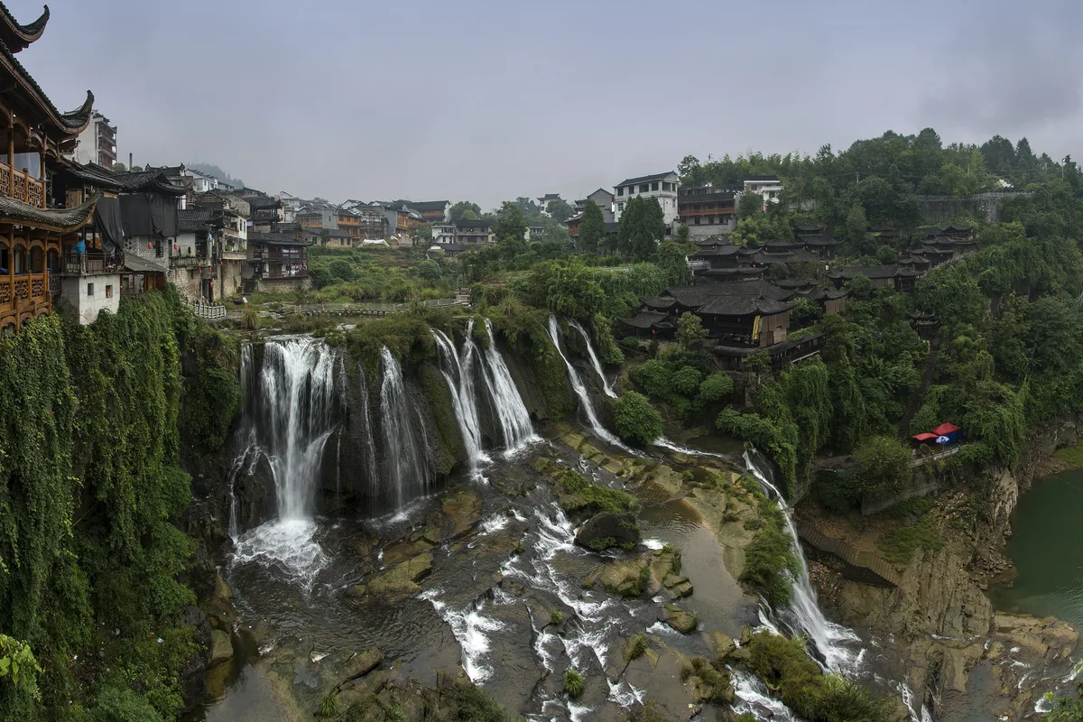 La cascada de Furong en dos niveles cayendo por la garganta, con casas sobre pilotes a la izquierda y el casco antiguo construido a lo largo del acantilado