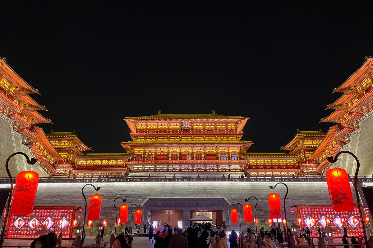 The floodlit gate tower of Yingtianmen in Luoyang at night, a three-tier Tang-style roof in gold light above a stone base, with crowds and red lanterns in the foreground (Photo: Windmemories via Wikimedia Commons)