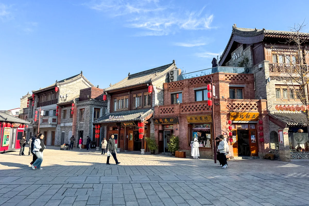 Une rangée de façades traditionnelles en brique grise avec lanternes rouges et fenêtres à cadre de bois sculpté sur une place pavée de la ville ancienne de Luoyi à Luoyang, quelques visiteurs en costume d'époque passant devant (Photo: Windmemories via Wikimedia Commons)