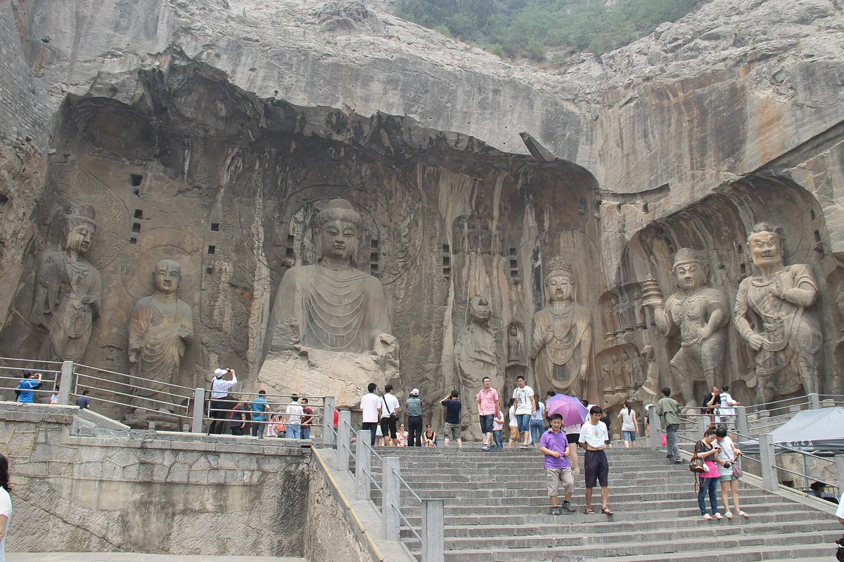 O buda colossal Vairocana e as figuras que o acompanham esculpidos no penhasco do templo Fengxian, nas grutas de Longmen, com visitantes nas escadas abaixo (Photo: Gary Todd via Wikimedia Commons)