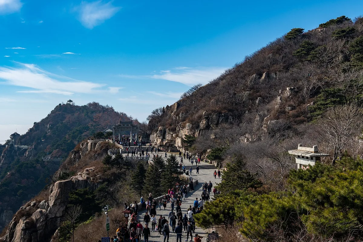 Visitors on the stone path along Mount Tai's summit ridge, with a temple gateway ahead and pine trees under a clear sky (Photo: xiquinhosilva via Wikimedia Commons)