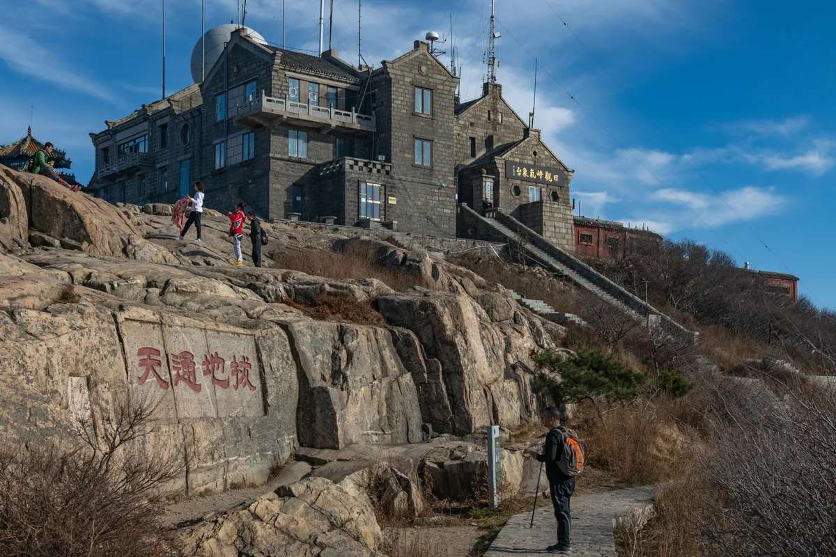 Los edificios de piedra de la estación meteorológica en la cresta de la cima del monte Tai, con la inscripción roja Badi Tongtian tallada en la roca y un caminante en el sendero de piedra
