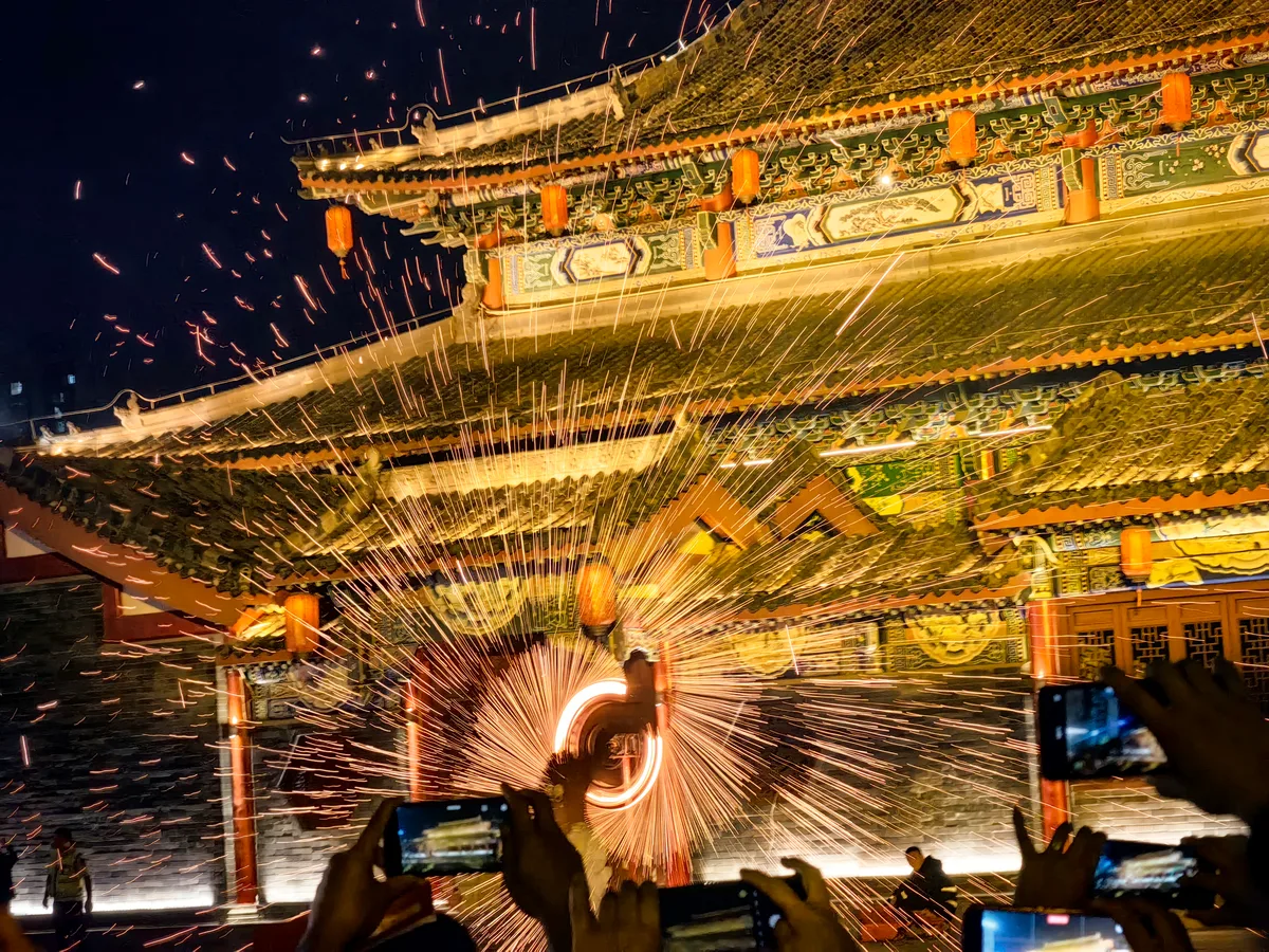 Hierro fundido lanzado contra la fachada tallada de un templo chino por la noche durante un espectáculo de flor de hierro, con el público grabando con el móvil (Photo: Auongkinghe via Wikimedia Commons)