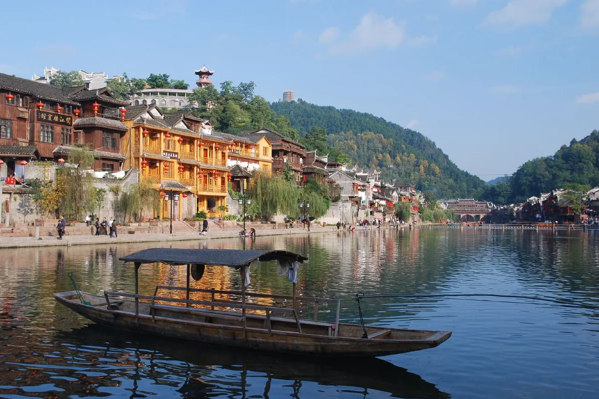 Una barca de madera amarrada en el río Tuojiang en Fenghuang, con las casas sobre pilotes y sus faroles rojos alineados en la orilla opuesta y colinas boscosas al fondo
