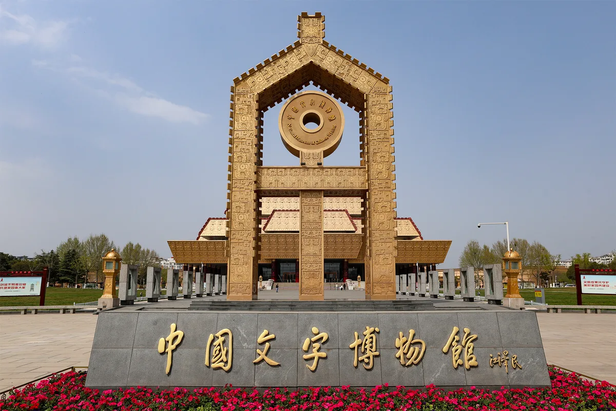 O pórtico do Museu da Escrita Chinesa em Anyang, uma estrutura dourada feita de formas de caracteres empilhadas, com o nome do museu gravado numa laje de pedra à frente (Photo: Windmemories via Wikimedia Commons)