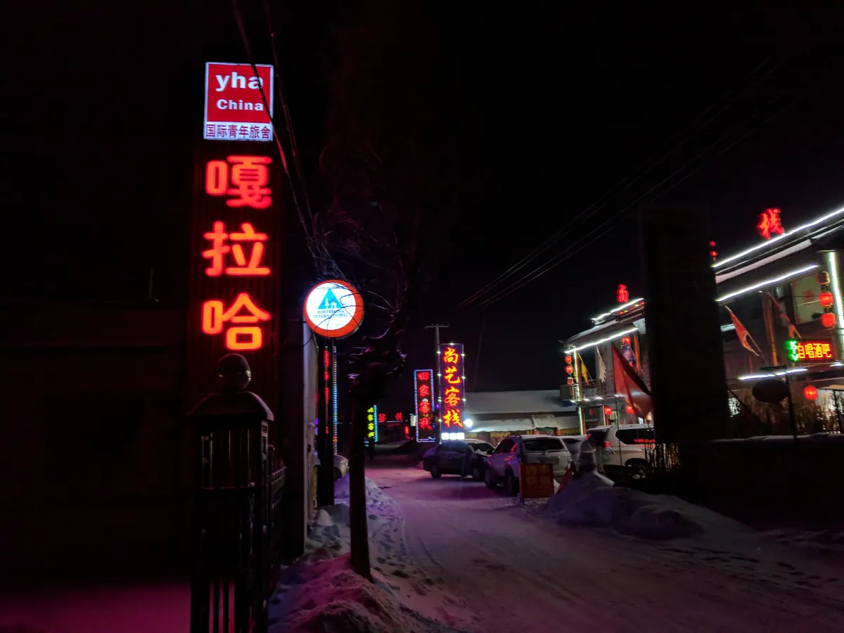 A youth hostel with a red neon sign on a snowy street in Jilin at night, Chinese shopfronts and parked cars alongside (Photo: Enming Yan via Wikimedia Commons)