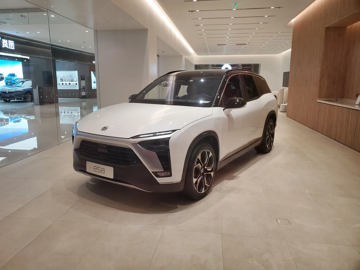 A white NIO ES8 displayed inside a NIO Space showroom, with a second car brand's showroom visible through the glass partition next door (Photo: 源義信 via Wikimedia Commons)