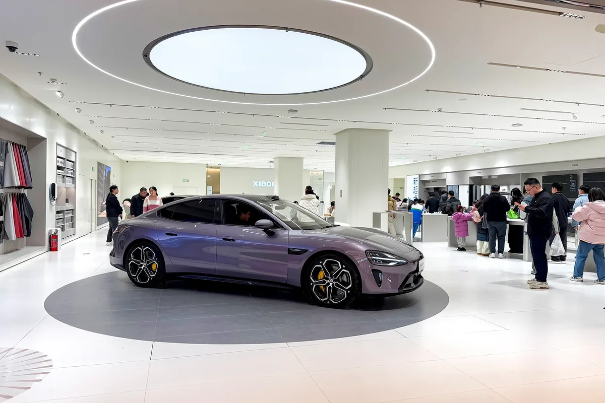A purple Xiaomi SU7 on a circular display platform inside a Xiaomi store, with shoppers browsing phones and home appliances around it (Photo: Windmemories via Wikimedia Commons)