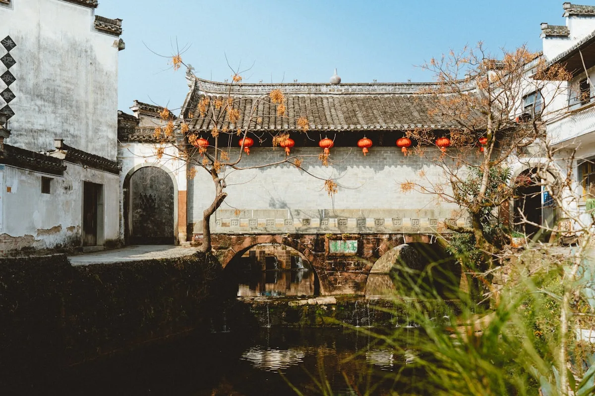 A Huizhou village stone bridge over a canal, white walls and grey tiled roofs with red lanterns along the water