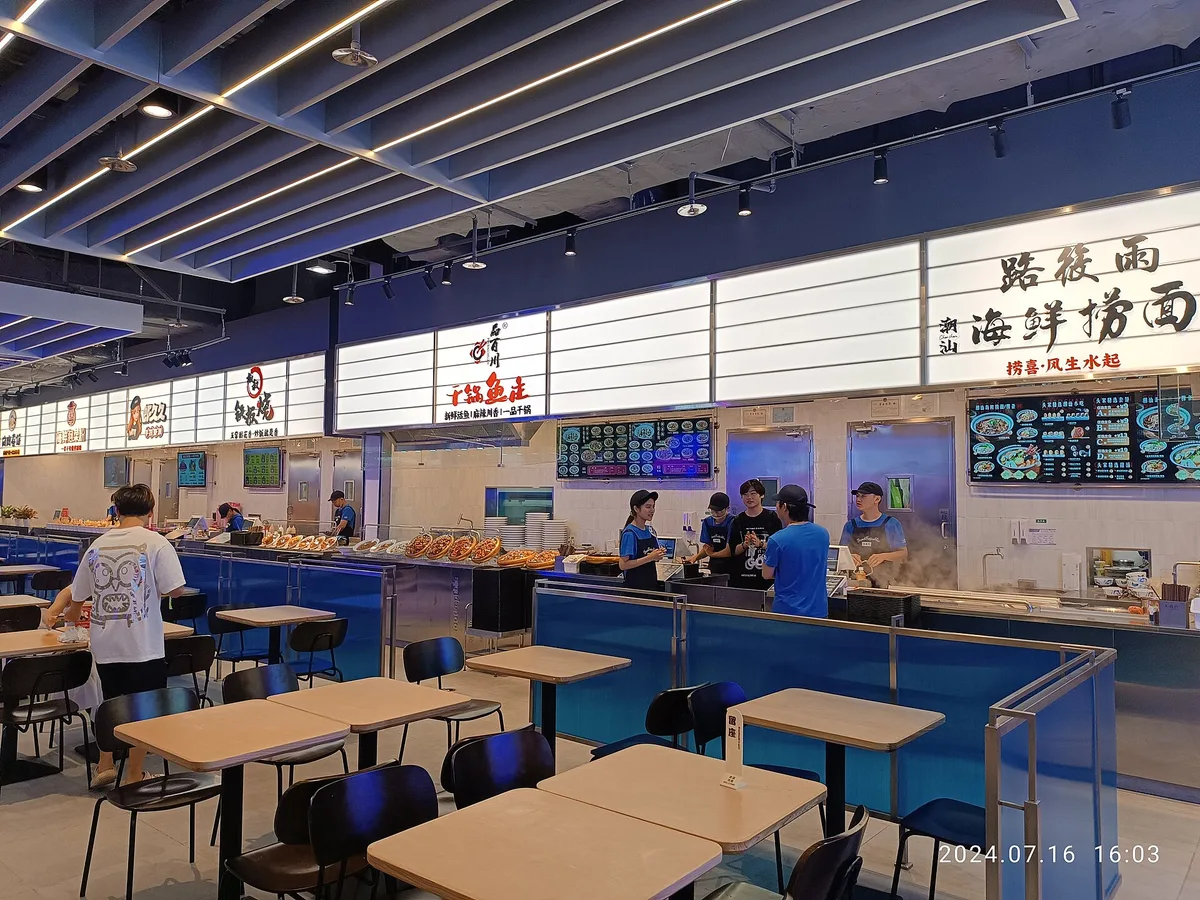 A food court inside a Shenzhen shopping mall, with staff working behind a row of lit food counters and diners seated at tables in front (Photo: ZHONG 82035 WANGZ via Wikimedia Commons)
