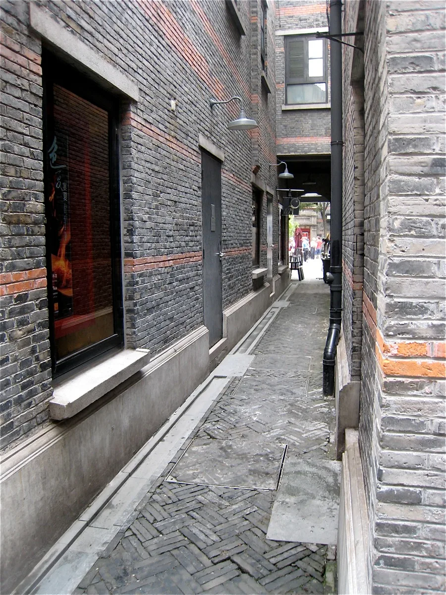 Uma viela estreita de tijolos no bairro shikumen de Xangai, com uma vitrine à esquerda e uma passagem coberta ao fundo (Photo: cogdogblog via Wikimedia Commons)