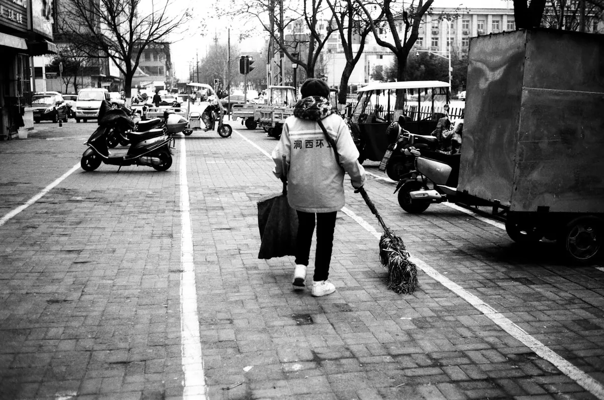 A street cleaner in a bright work vest sweeping a Chinese city street, scooters parked along the pavement