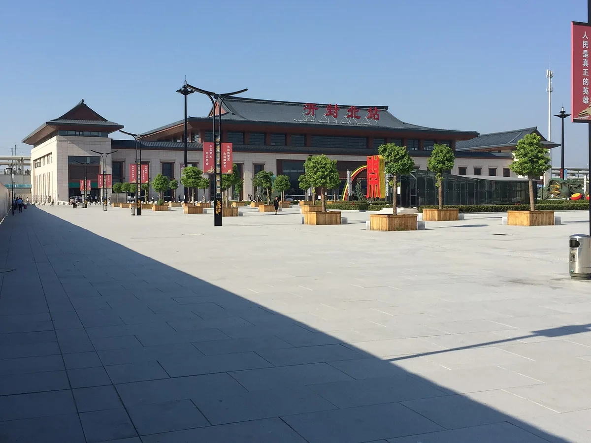 O prédio da estação de Kaifeng Norte, um galpão de telhados cinza escalonados com o nome da estação em caracteres vermelhos, vazio sob o calor do meio-dia (Photo: Kestreltail via Wikimedia Commons)