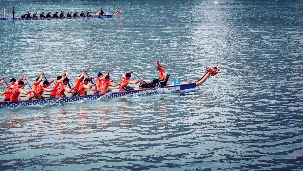 A dragon boat crew in orange rows hard down the river, the carved dragon head at the prow cutting the water