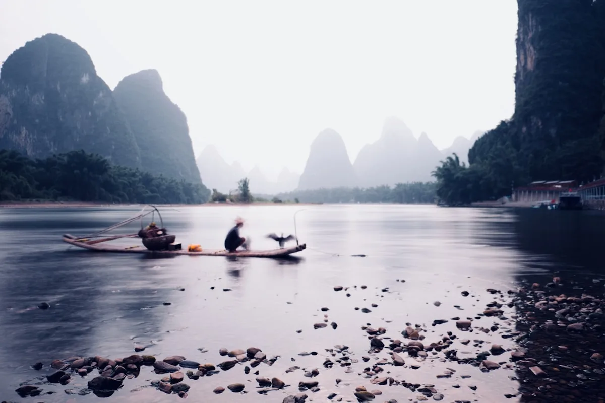 A fisherman standing on his bamboo raft out on the Li River, karst peaks and morning mist rising behind him near Yangshuo