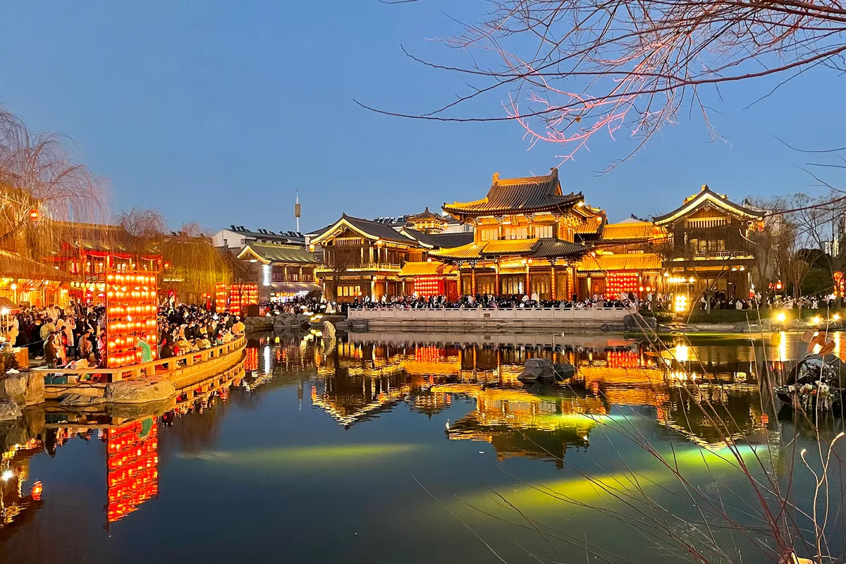 A cidade antiga de Luoyi ao anoitecer, um pavilhão iluminado de vários níveis junto a um lago com lanternas refletidas na água e multidão na margem oposta (Photo: Windmemories via Wikimedia Commons)