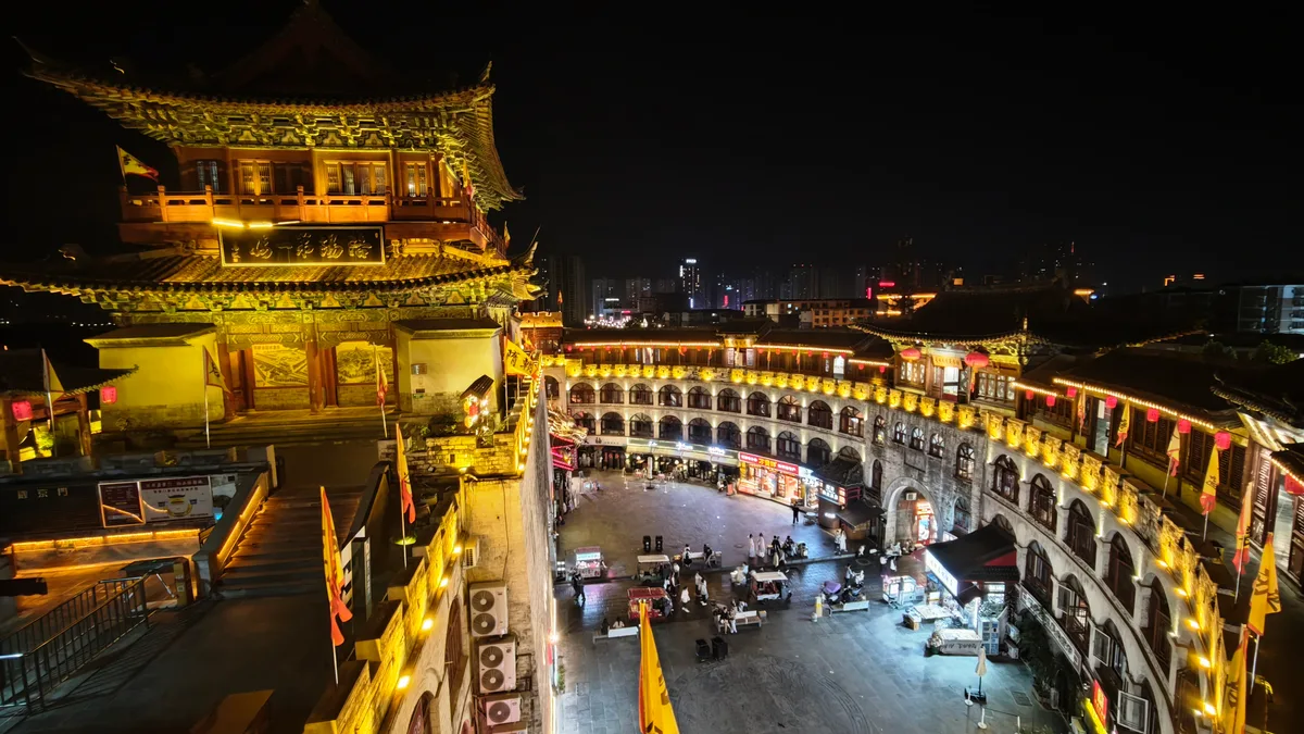 La porte de Lijing à Luoyang de nuit vue d'en haut, sa tour illuminée au-dessus d'une place de marché entourée de bâtiments à arcades, avec les étals et la foule en contrebas (Photo: Renek78 via Wikimedia Commons)