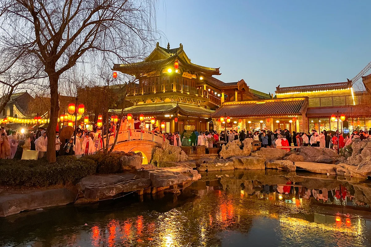 La ville ancienne de Luoyi à Luoyang au crépuscule, bâtiments de bois éclairés et lanternes rouges reflétés dans un bassin, avec une foule de visiteurs dont plusieurs en hanfu sur la rive opposée (Photo: Windmemories via Wikimedia Commons)