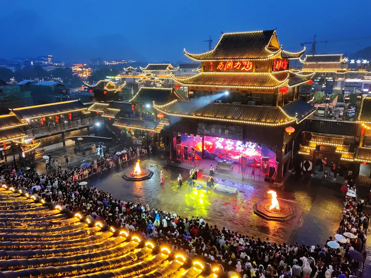 Multitud reunida alrededor de las hogueras en el patio del teatro Charming Xiangxi de noche, frente al escenario exterior iluminado