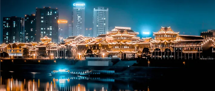 El complejo de la ciudad antigua de Dayong en Zhangjiajie iluminado de noche, con sus tejados escalonados reflejados en el río bajo los rascacielos de la ciudad moderna