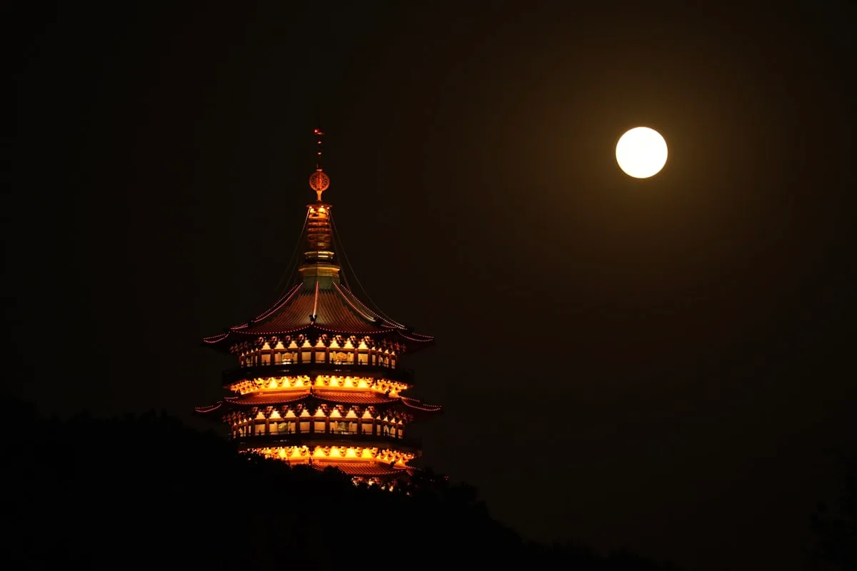 The Leifeng Pagoda on the shore of Hangzhou's West Lake lit up at night beside a full moon, the classic Mid-Autumn view
