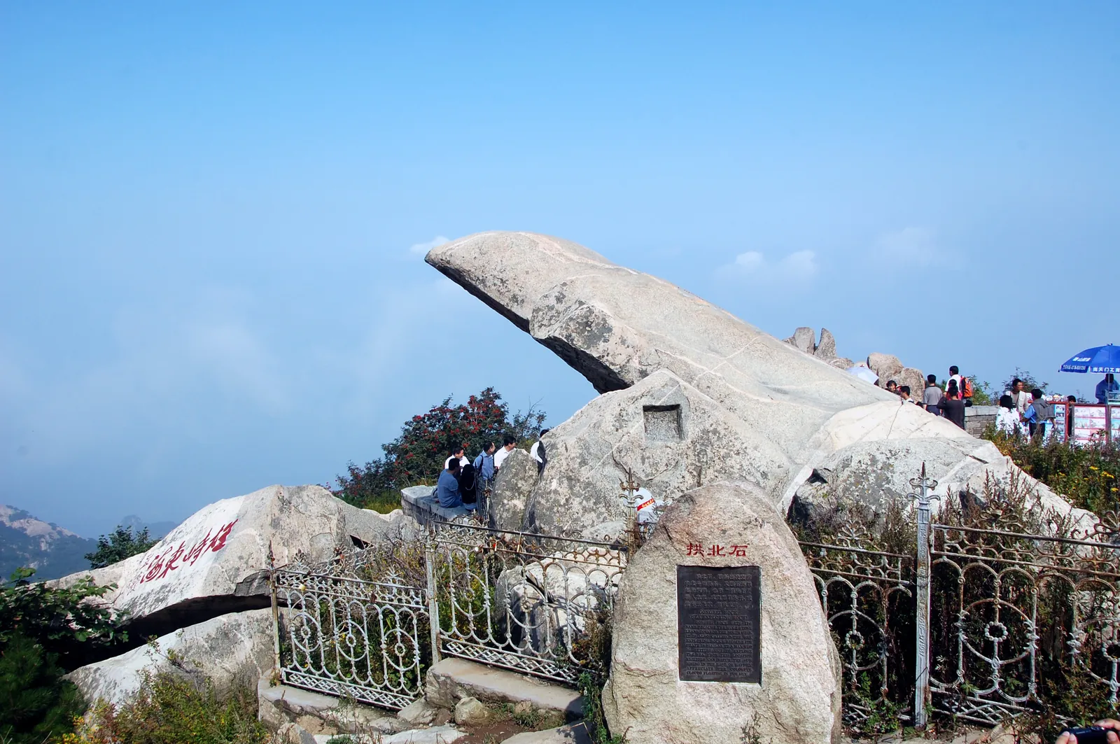 La roca Gongbei en voladizo sobre la cresta de la cima del monte Tai, detrás de una verja baja y una placa de piedra, con visitantes reunidos en la barandilla más allá (Foto: kanegen vía Wikimedia Commons)