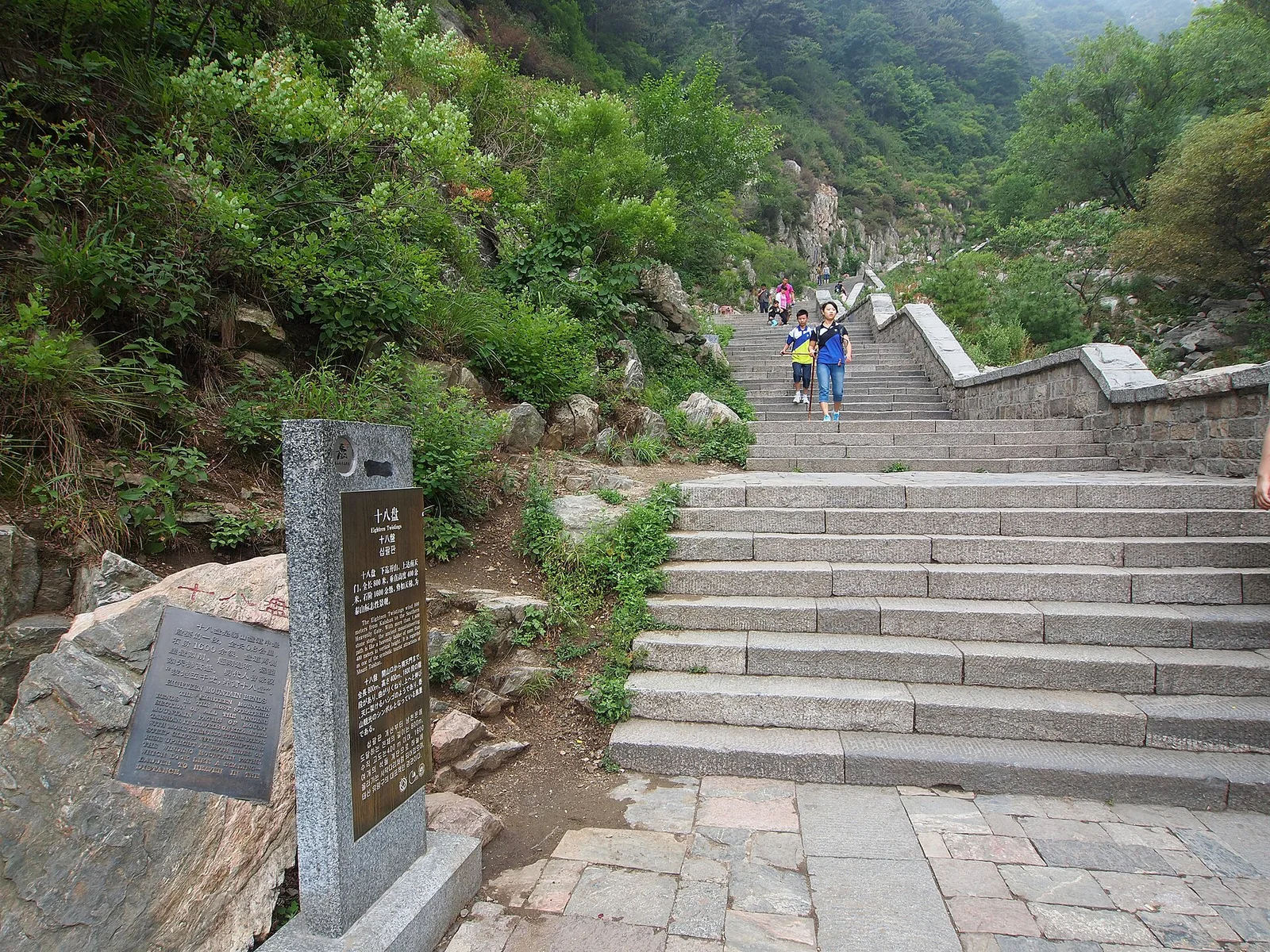 The granite marker at the foot of the Eighteen Bends on Mount Tai, its carved plaque giving the step count, with the staircase climbing into green hillside behind it