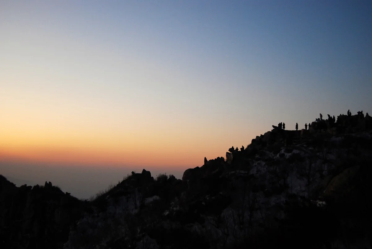 A crowd of visitors silhouetted on Mount Tai's summit ridge before sunrise, with the sky glowing orange over a hazy valley (Photo: shizhao via Wikimedia Commons)