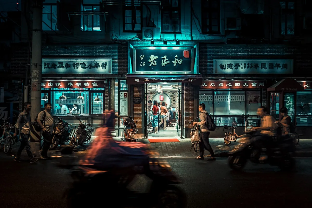 People and e-bikes passing a brightly lit restaurant entrance on a Shanghai street at night