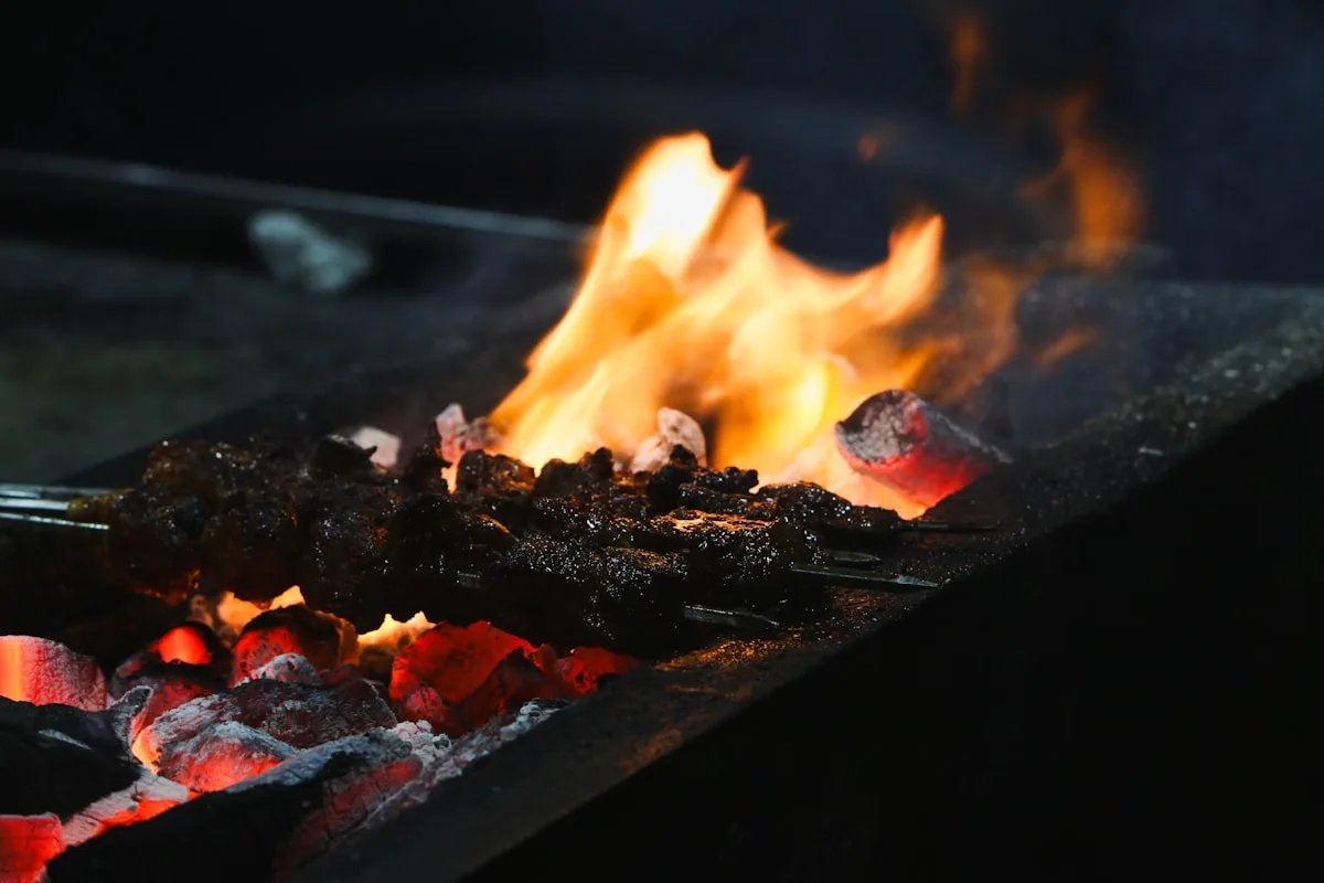 Charred meat skewers cooking over glowing charcoal with small flames rising between the grill bars