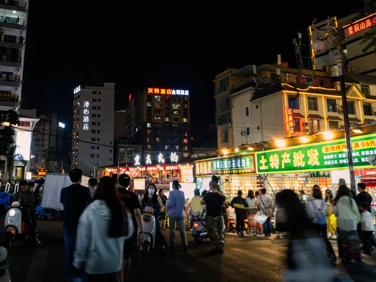 Crowds walking past brightly lit shops and food stalls on a Chinese night-market street in the evening