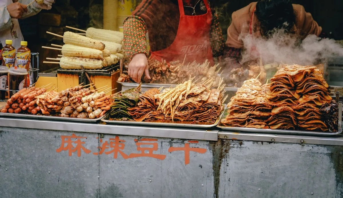 A night-market vendor in a red apron cooking at a stall piled with skewers and snacks while steam rises