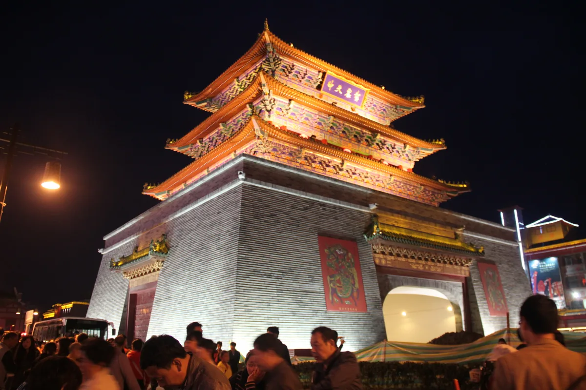 La tour du Tambour de Kaifeng illuminée la nuit, son passage voûté ouvert à la circulation, avec une foule et des taxis sur la place devant (Photo: Gary Todd via Wikimedia Commons)