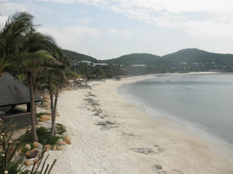 A quiet stretch of white sand at Yalong Bay, with coconut palms and resort buildings behind and green hills across the water (Photo: Kusafiri via Wikimedia Commons)