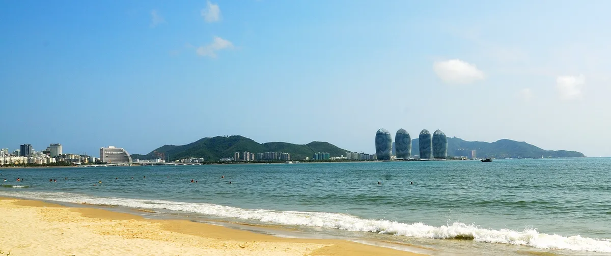 The beach at Sanya Bay with the Phoenix Island towers on the horizon and swimmers in the water (Photo: Anna Frodesiak via Wikimedia Commons)