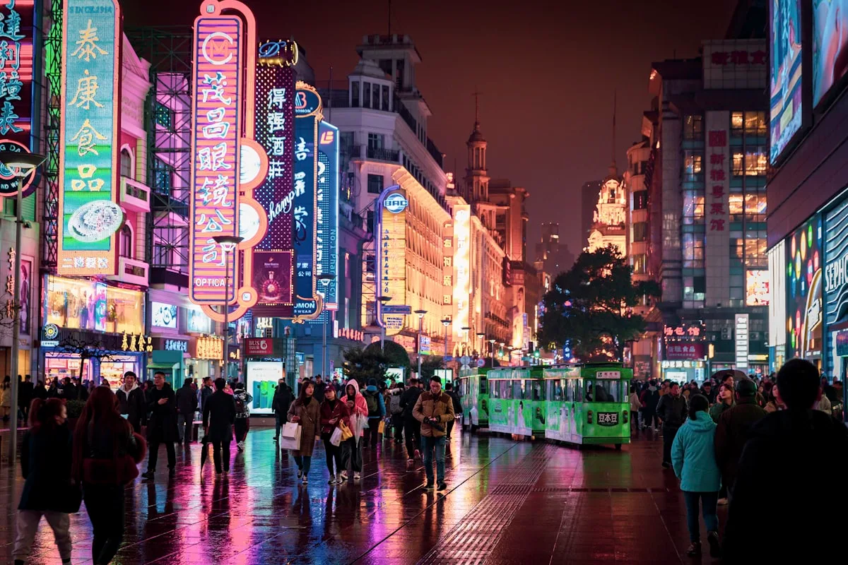 A neon-lit shopping street in Shanghai crowded with evening pedestrians after rain, showing a busy late-night city center