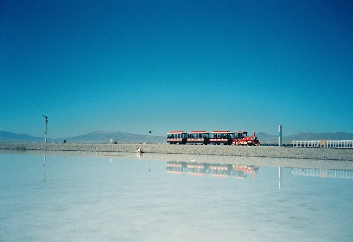 A red train crossing the white salt flats of Chaka Salt Lake, its reflection still on the mirror-like water beneath a blue sky