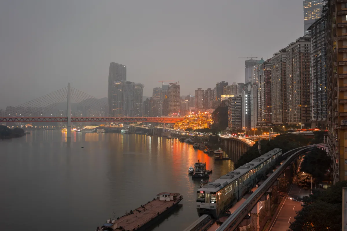 A Chongqing monorail running on an elevated track beside the Yangtze River at night, with the Qiansimen Bridge and the lit-up Hongyadong stilt houses behind it (Photo: Kristoffer Trolle via Wikimedia Commons)