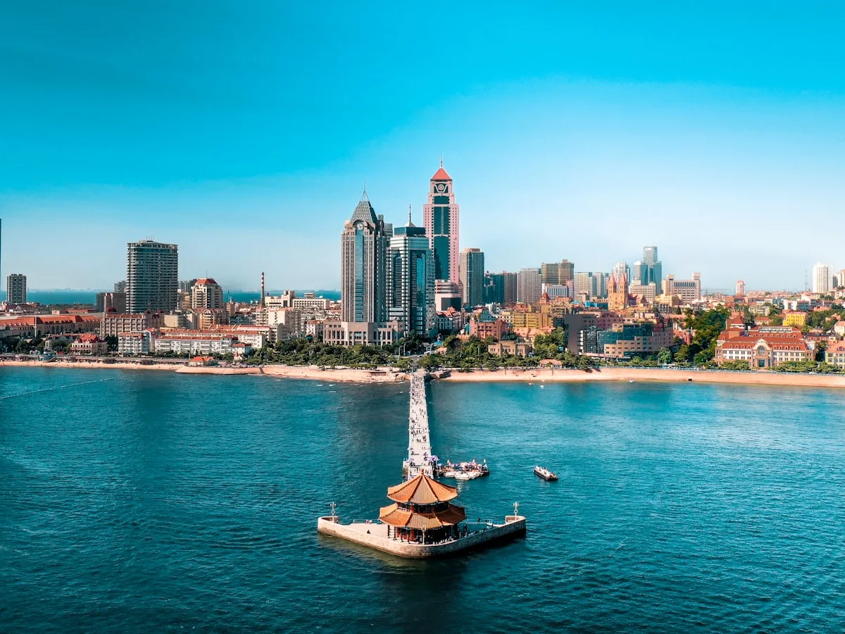 Aerial view of Qingdao's Zhanqiao pier and Chinese pavilion stretching into the sea beside red-roofed buildings and a modern skyline