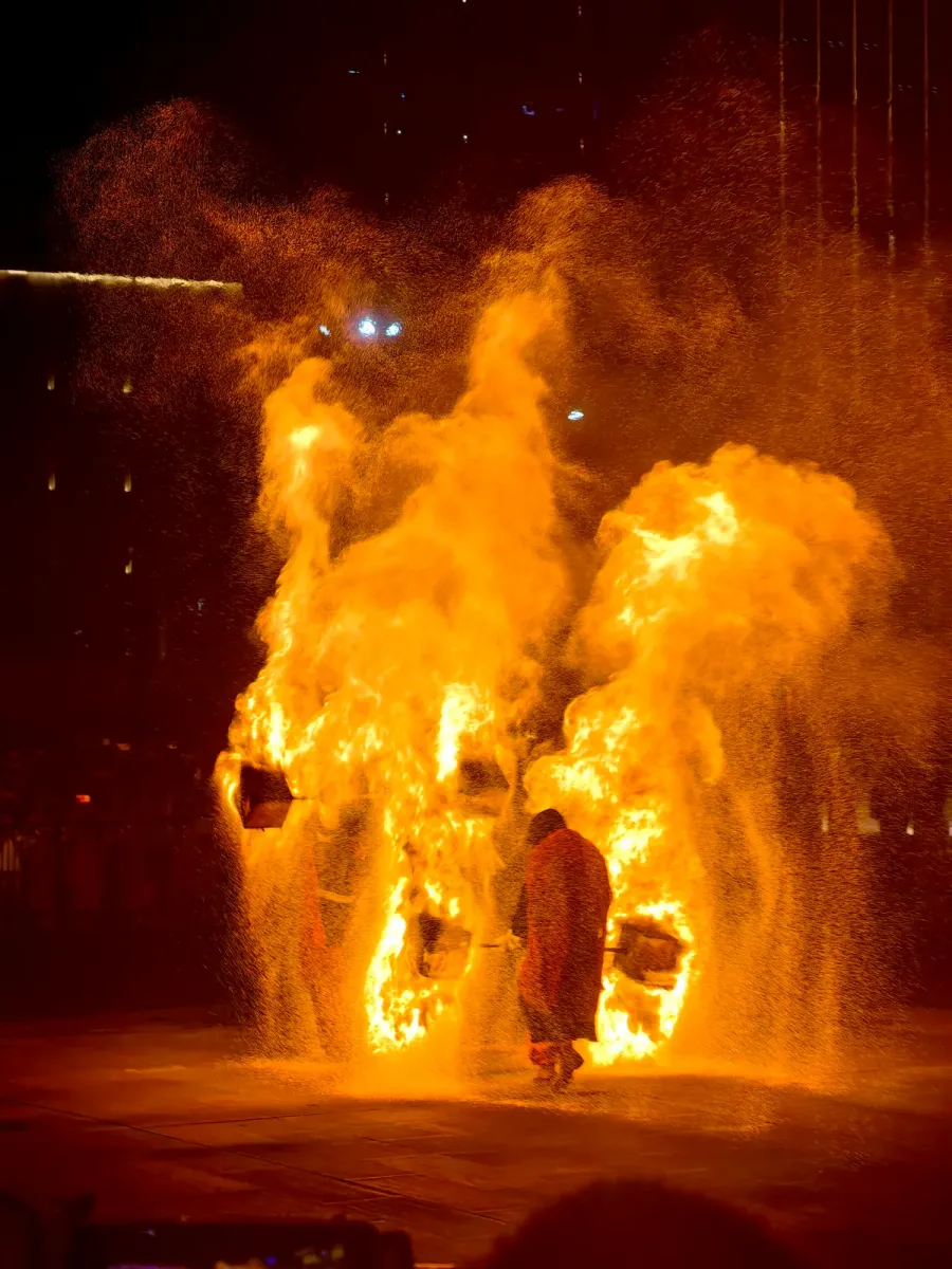 Un artista con túnica roja cruza una cortina de chispas durante un espectáculo nocturno de flor de hierro, con el público y las luces de la ciudad detrás (Photo: Auongkinghe via Wikimedia Commons)