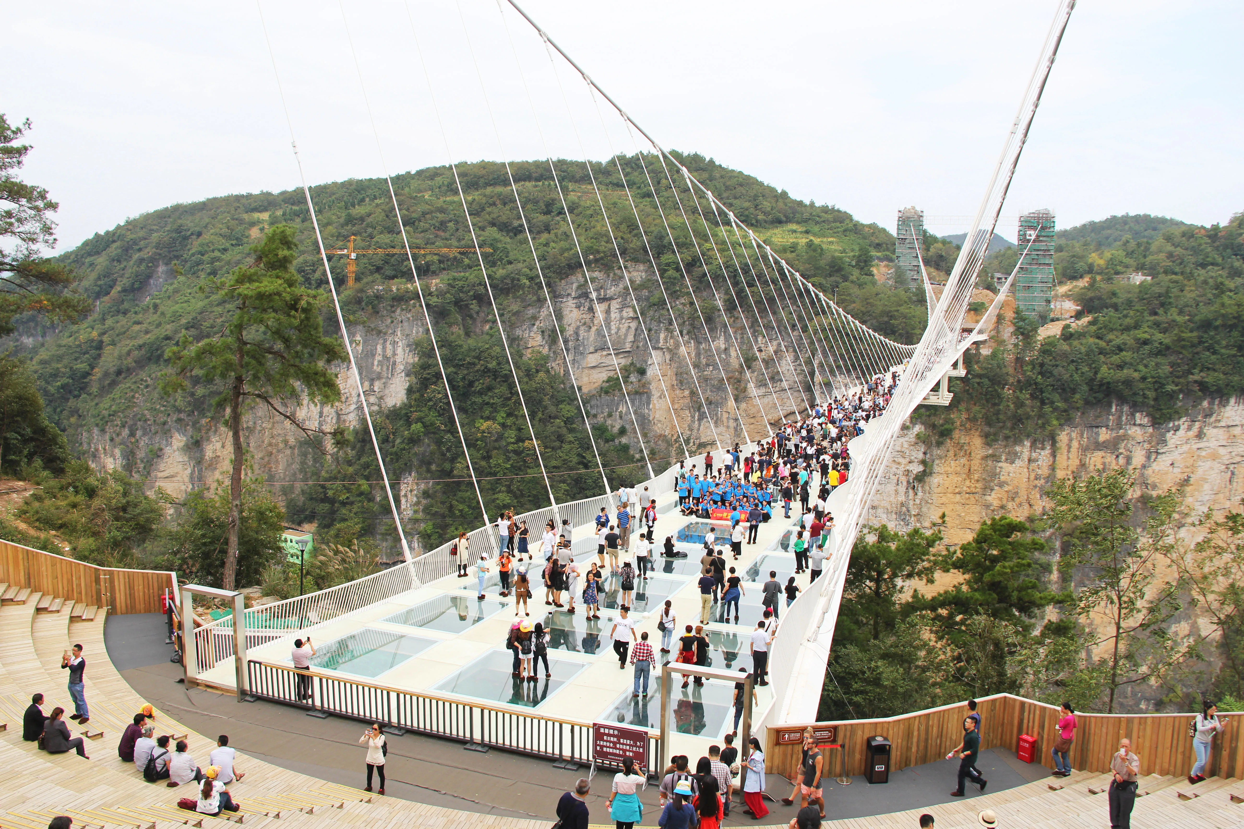 Uma multidão de visitantes atravessando o tabuleiro transparente, com os cabos estaiados e as paredes do cânion ao fundo