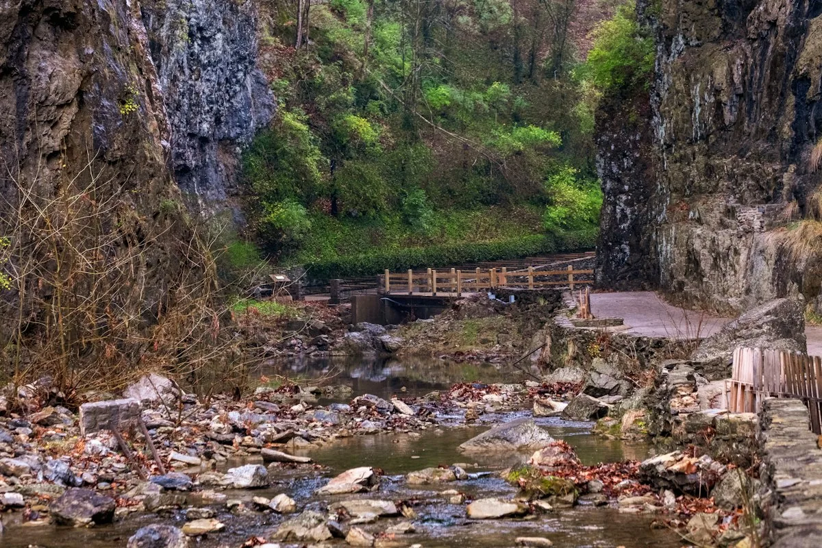 Uma passarela pavimentada e uma ponte de madeira cruzando um riacho pedregoso entre paredes altas de rocha, com folhas de outono ao longo da água