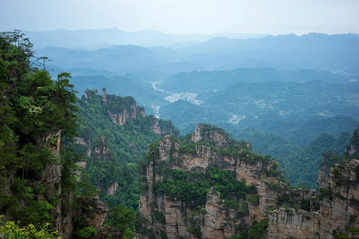 Layered sandstone pillars rising above a forested valley, with a small settlement on the valley floor inside Zhangjiajie National Forest Park