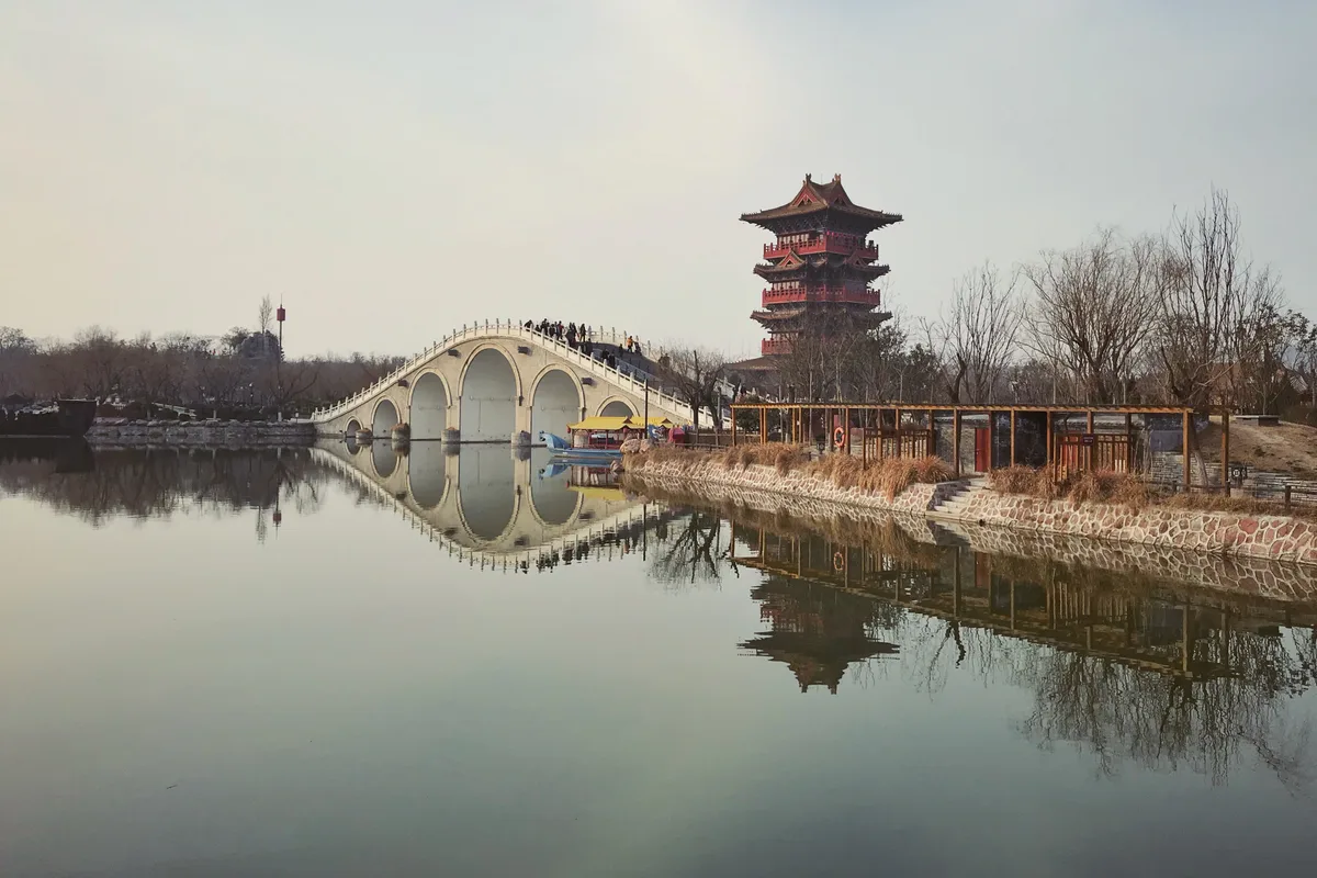 A stone arch bridge reflected in still water at Qingming Shanghe Garden in Kaifeng, with a multi-tiered wooden pavilion on the far bank (Photo: Windmemories via Wikimedia Commons)