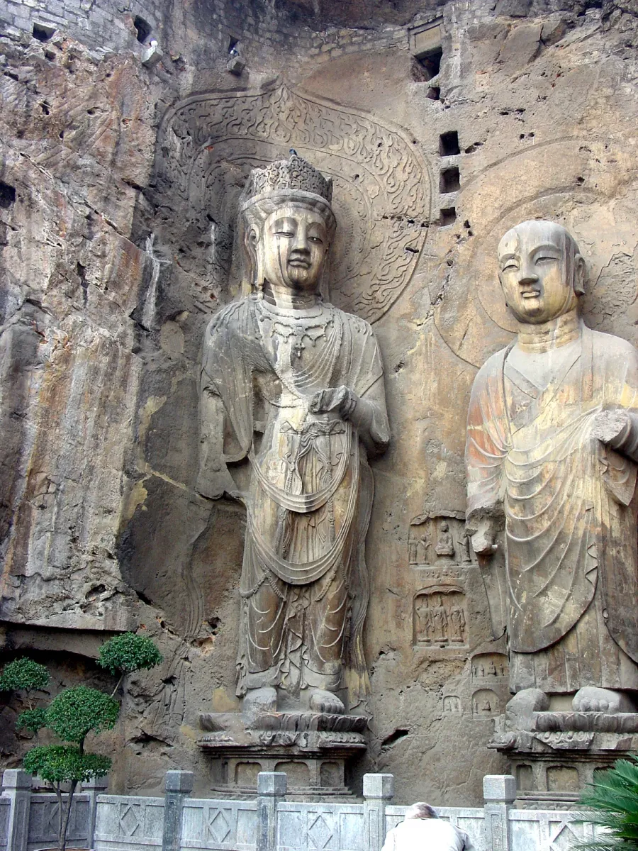 Two carved stone bodhisattva figures standing against the cliff face at the Longmen Grottoes near Luoyang, with smaller niches cut into the rock between them (Photo: G41rn8 via Wikimedia Commons)